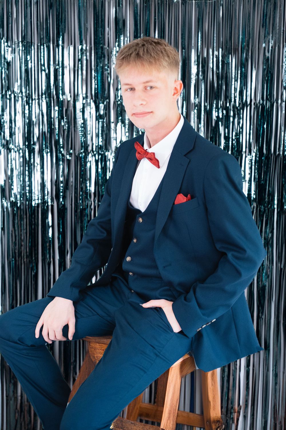 Dapper young man in navy suit with red bow tie poses on stool against silver backdrop.