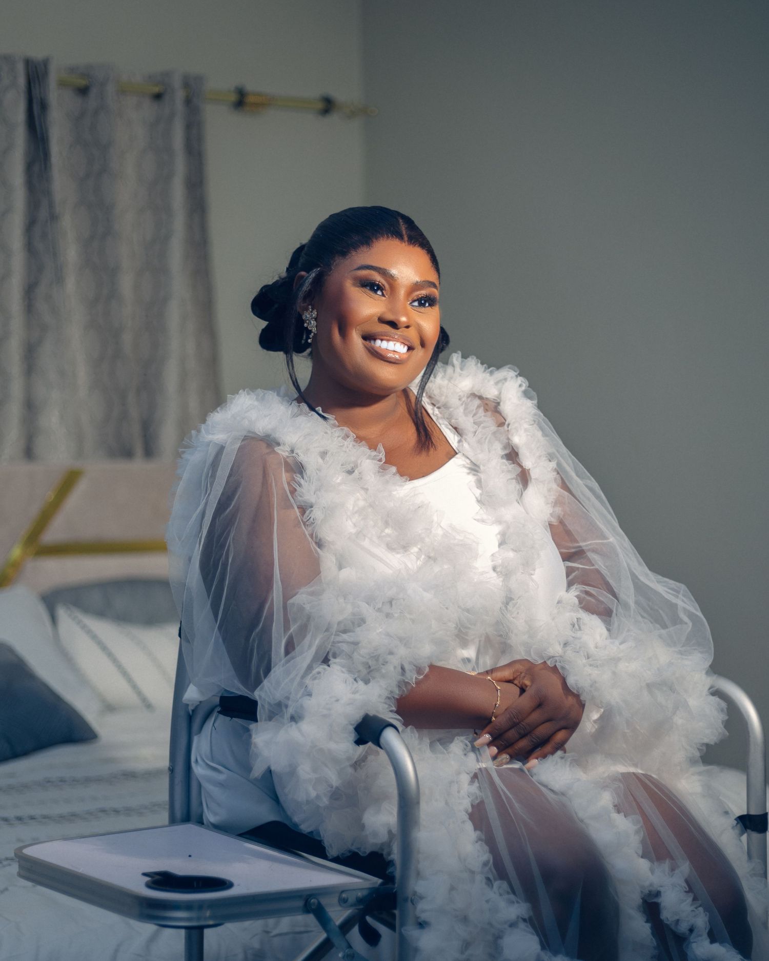 A smiling person wears an ethereal white gown with tulle and feather details while sitting in a bright interior setting.