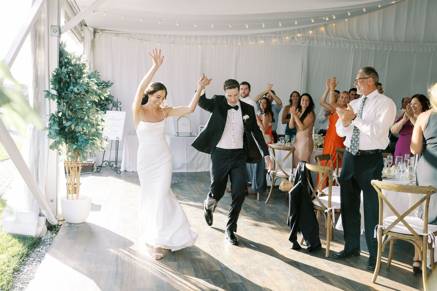 Wedding guests celebrate under a white tent decorated with string lights as people dance and enjoy the reception festivities.