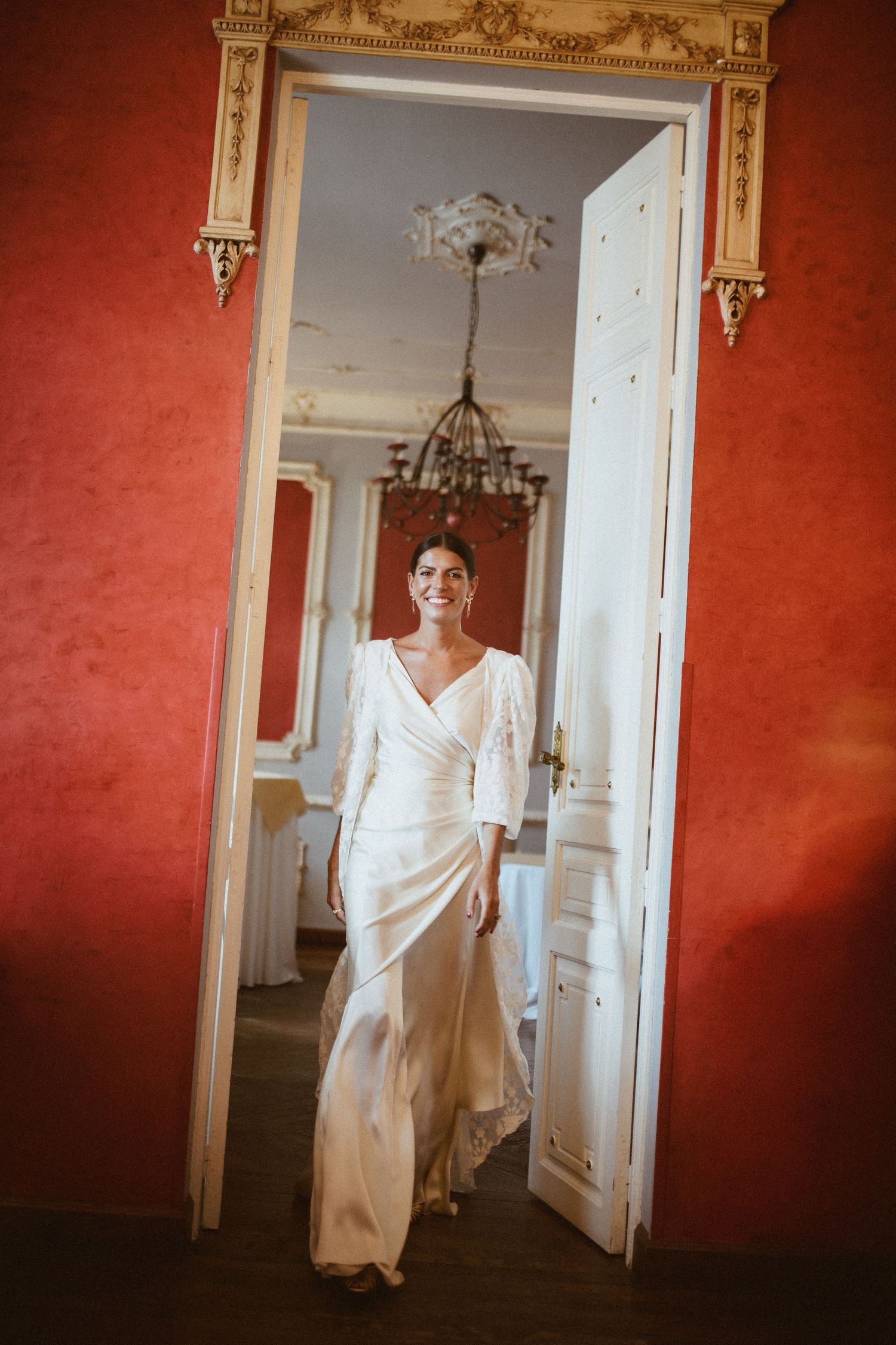 A bride in a flowing white dress poses elegantly through a doorway in an ornate room with red damask walls.