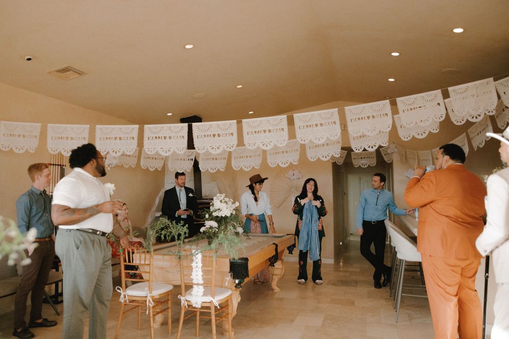 Indoor party scene with white decorative banners and people gathered in a warm-toned room with chairs and decorations.