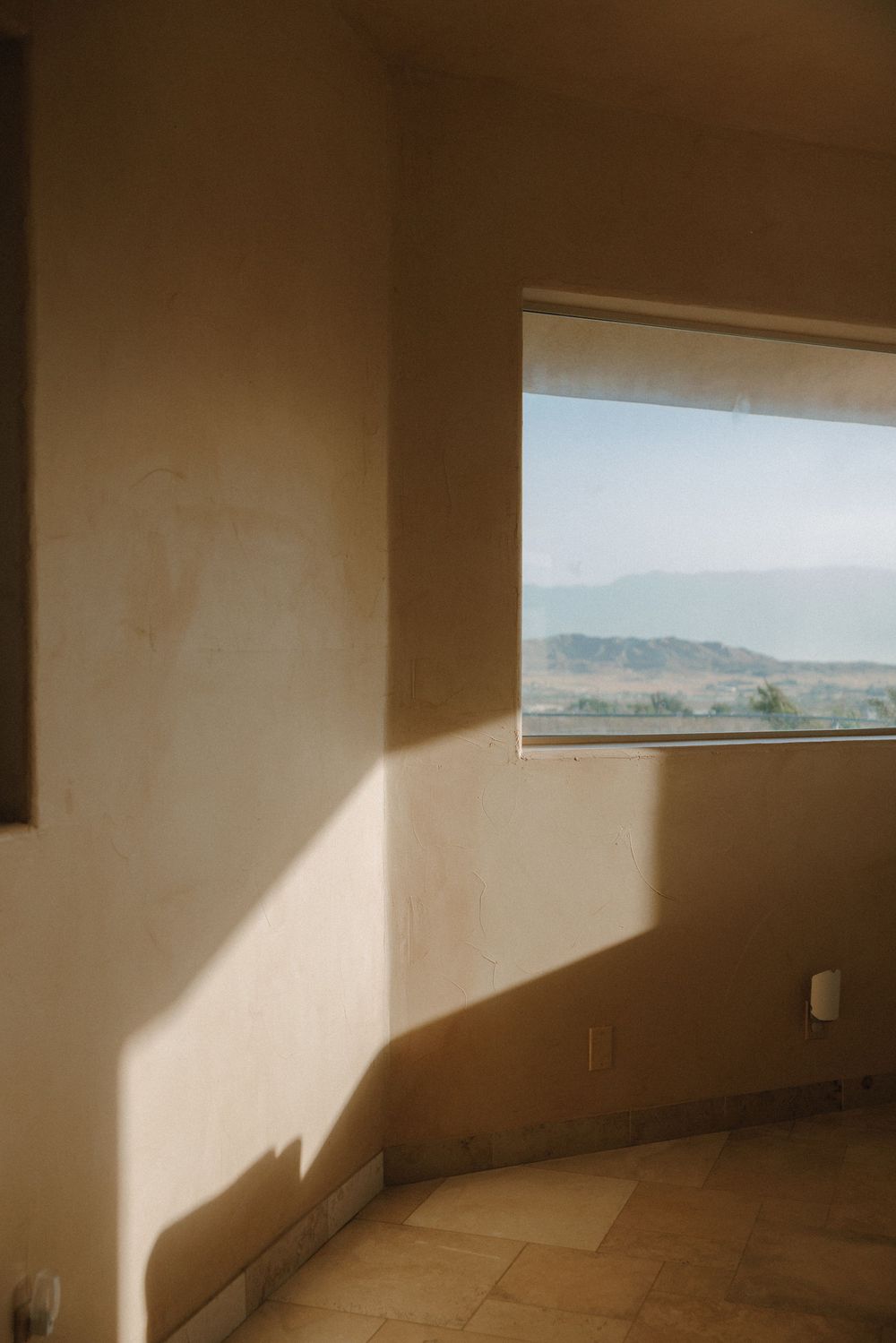 Sunlight casts dramatic shadows across an empty room with a window overlooking distant mountains and hills.