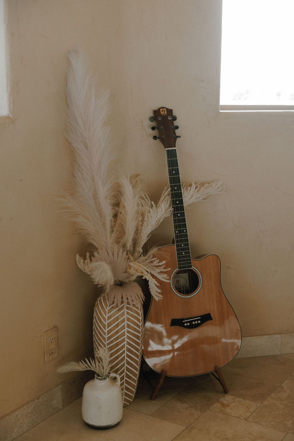 Acoustic guitar leans against beige wall with dried pampas grass in chevron patterned vase creating minimal home decor scene.