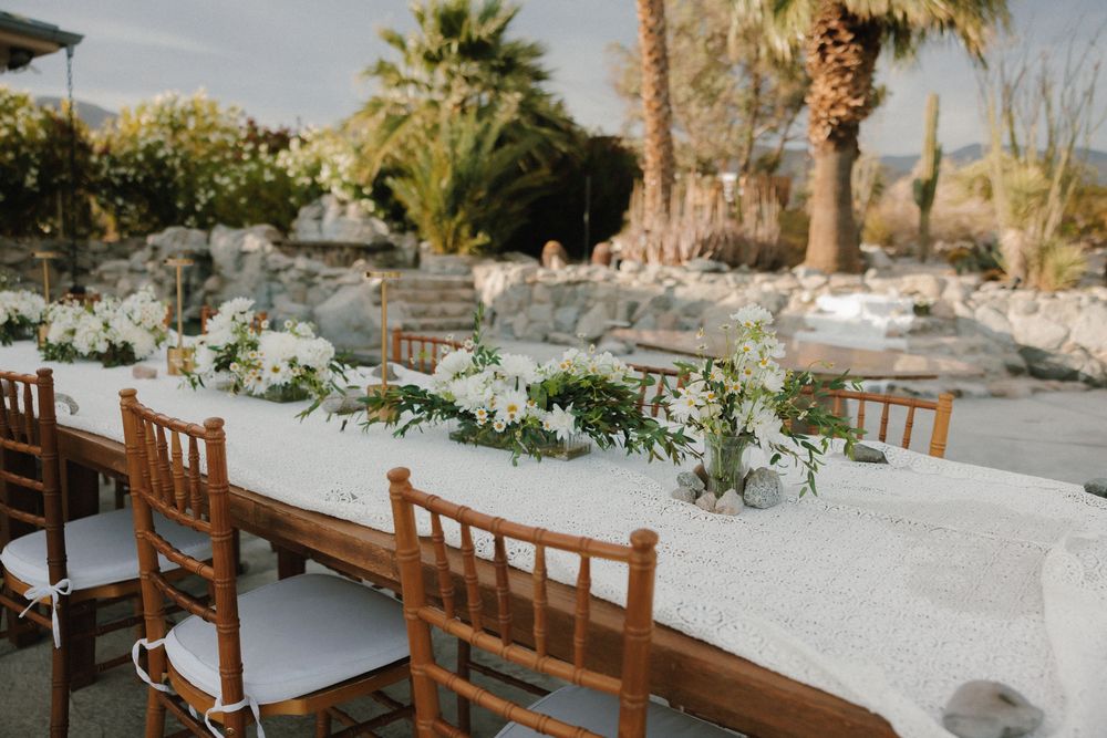 Elegant outdoor wedding reception table with wooden chairs and white linens set against a desert landscape with palm trees.