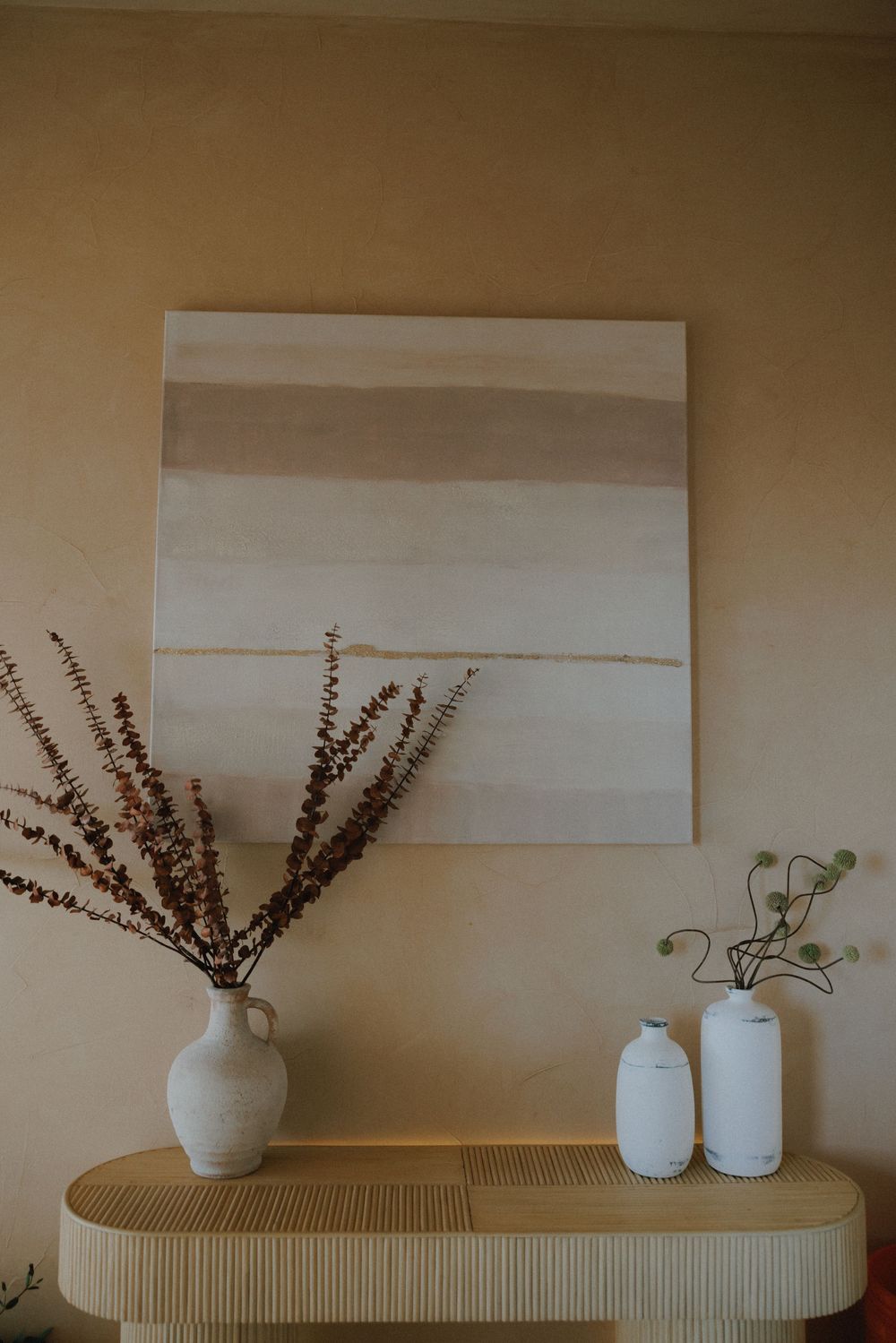 Minimalist home decor featuring neutral abstract art, dried flowers in a ceramic vase, and white jars on a curved wooden shelf.