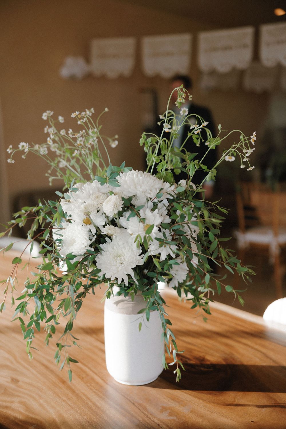 White floral centerpieces with greenery arranged on a wooden table in gentle natural light.