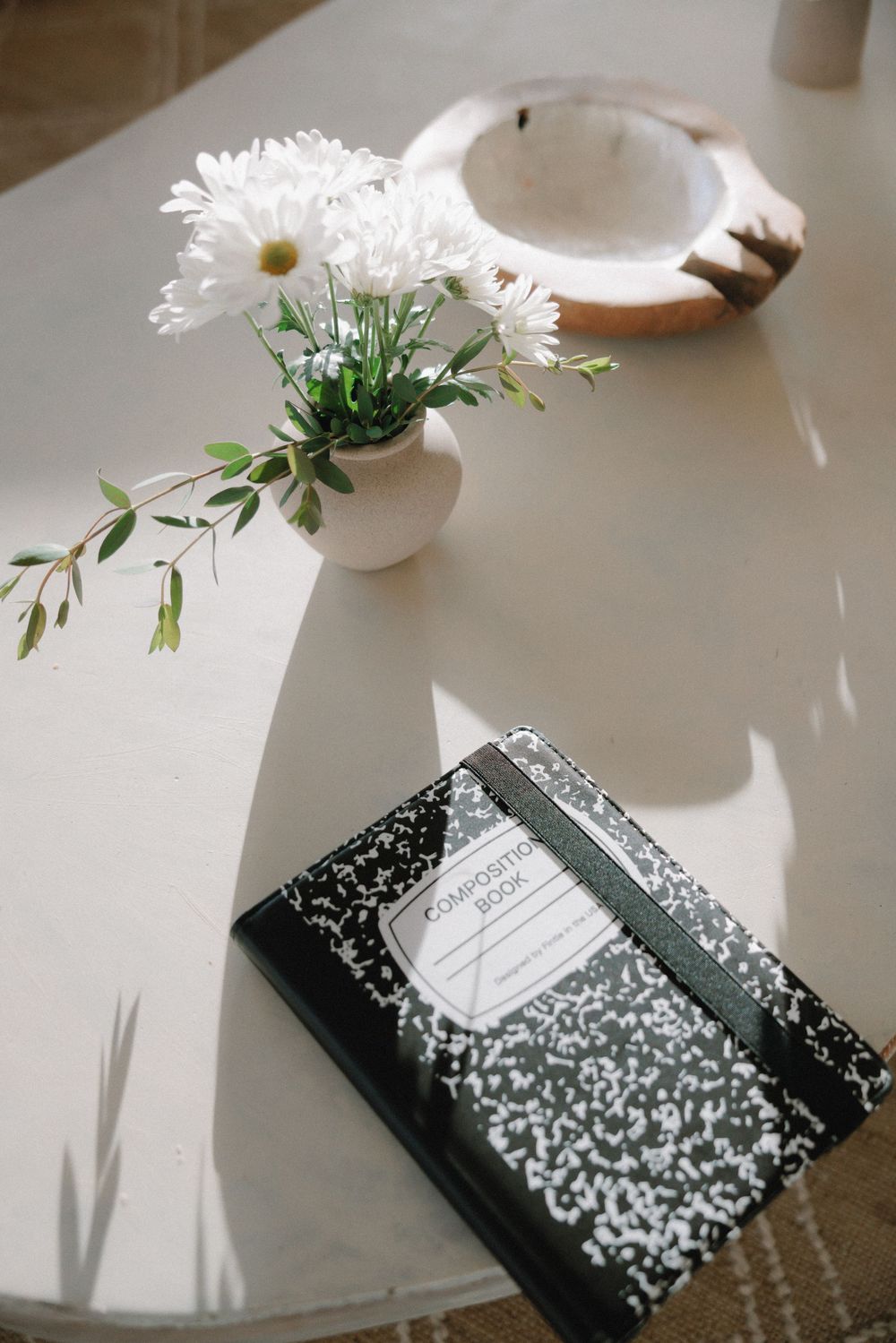 A composition notebook sits on a white surface next to a small vase of white daisies and greenery.