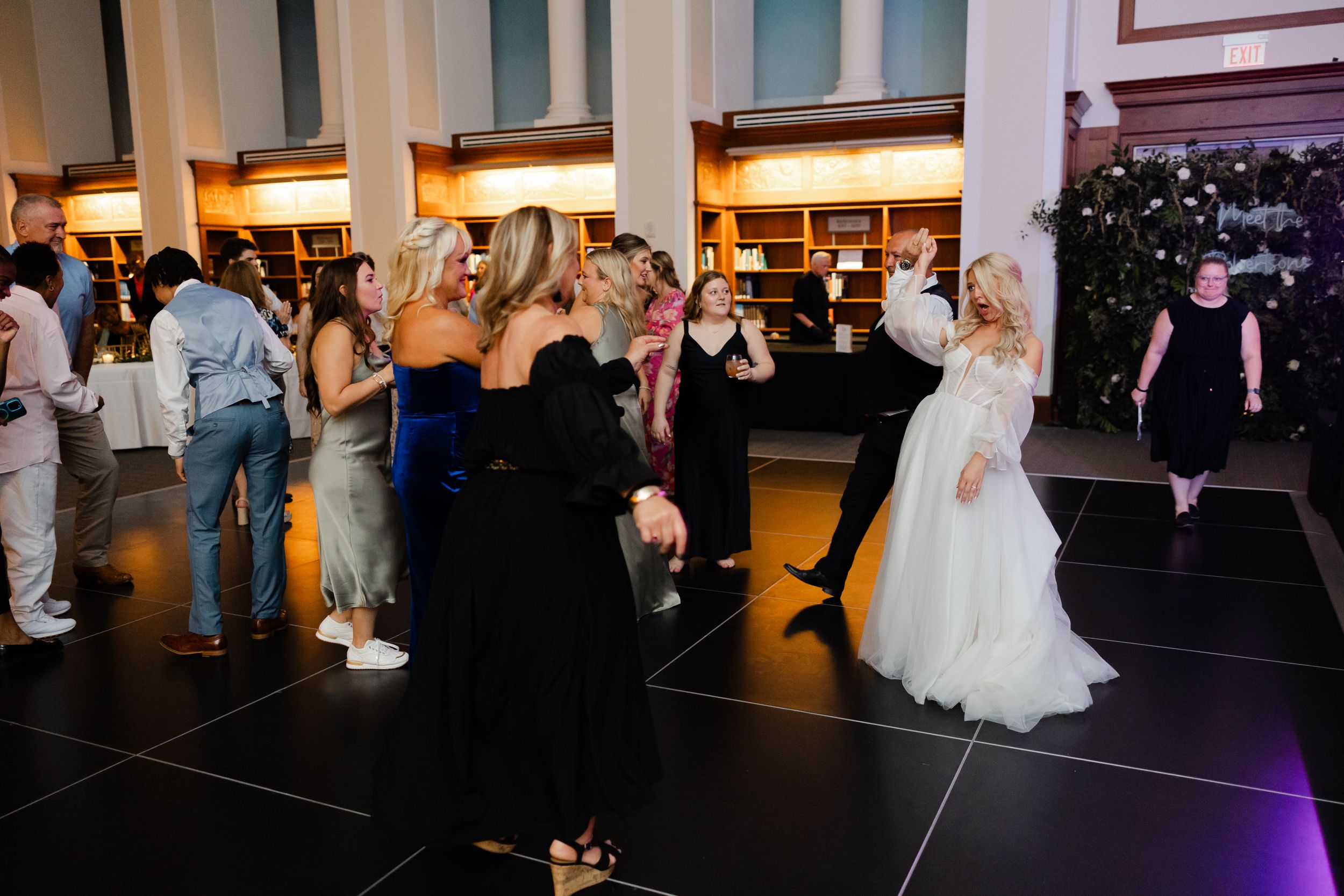 Wedding guests dance and celebrate together on a dark dance floor illuminated by warm lighting at an indoor reception.