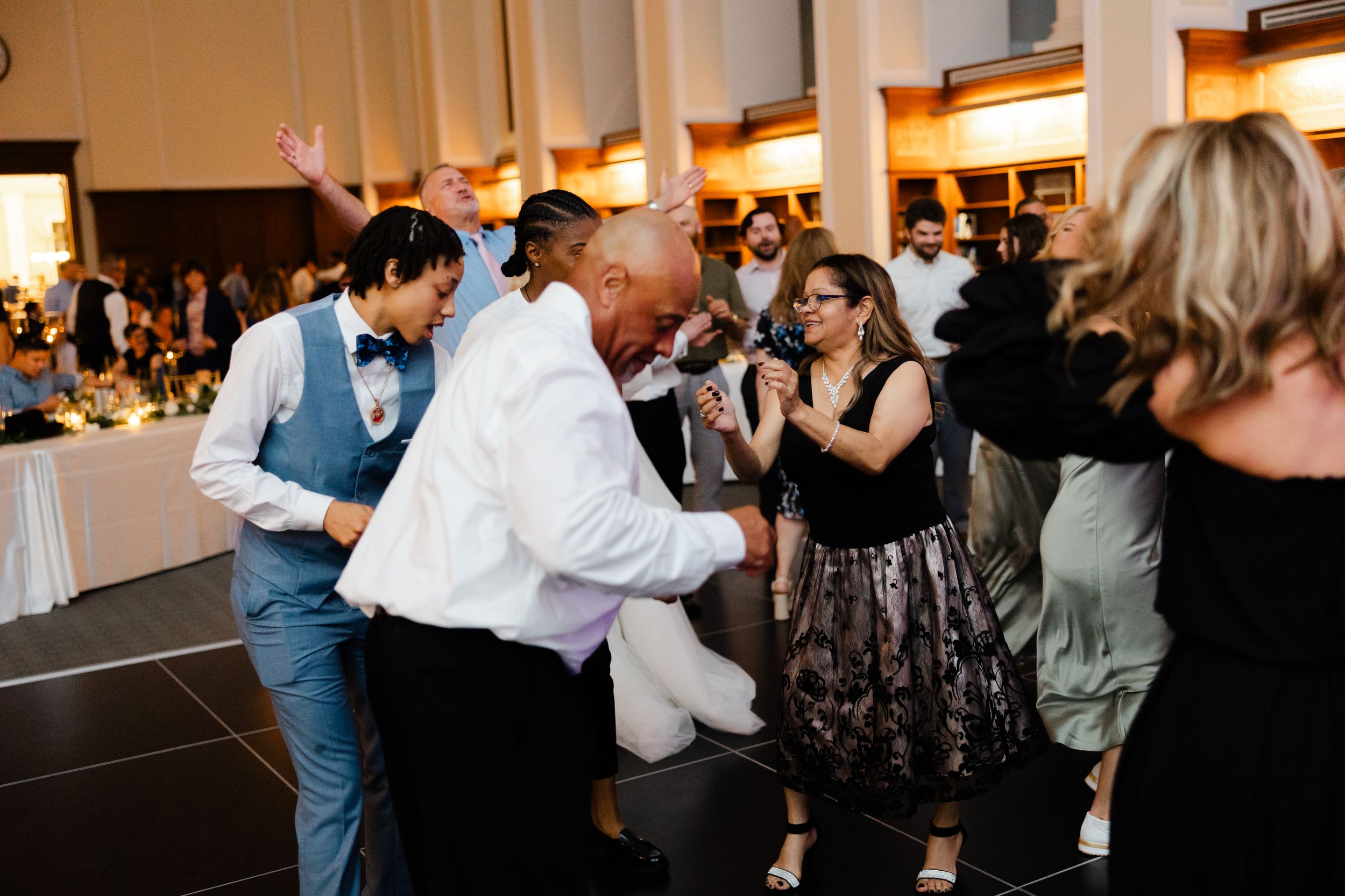 Wedding guests dance and celebrate together on a dark dance floor at an indoor reception venue with white pillars and lighting.