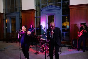 Live band performs on stage with singers and musicians under colorful purple lighting at night venue.