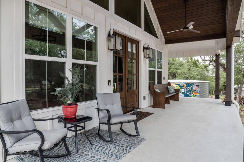 A spacious covered porch with gray rocking chairs and a seating area framed by large windows overlooks a tranquil backyard.