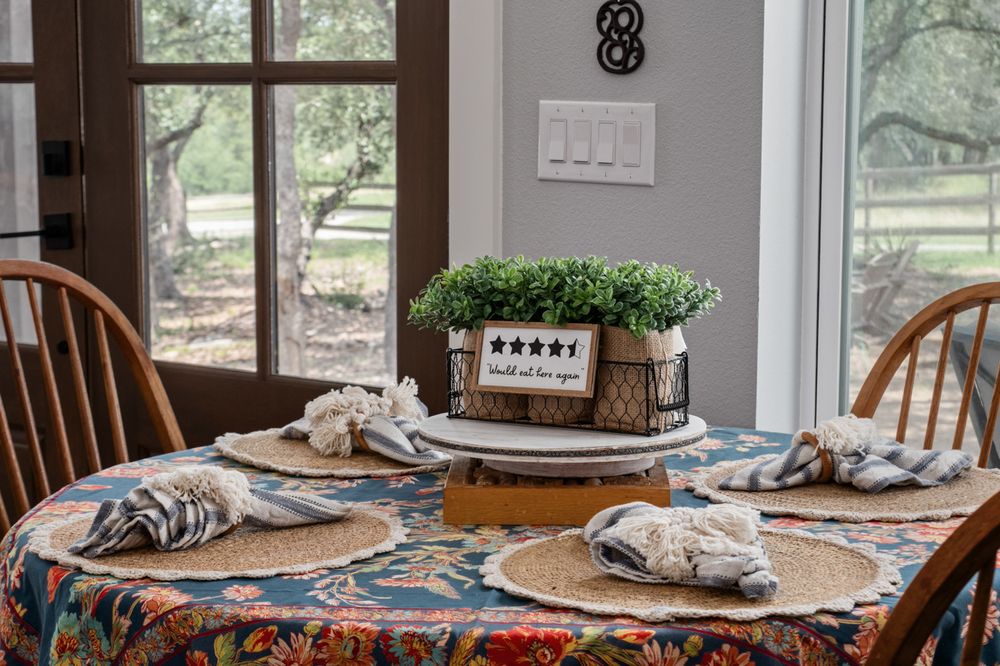 Rustic dining table with floral tablecloth, wooden chairs, and centerpiece box of fresh herbs near sunny windows.