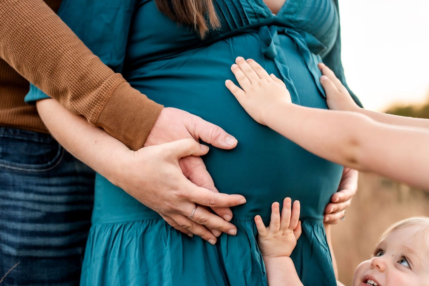 Gentle hands rest on a pregnant belly in teal dress against a soft background.