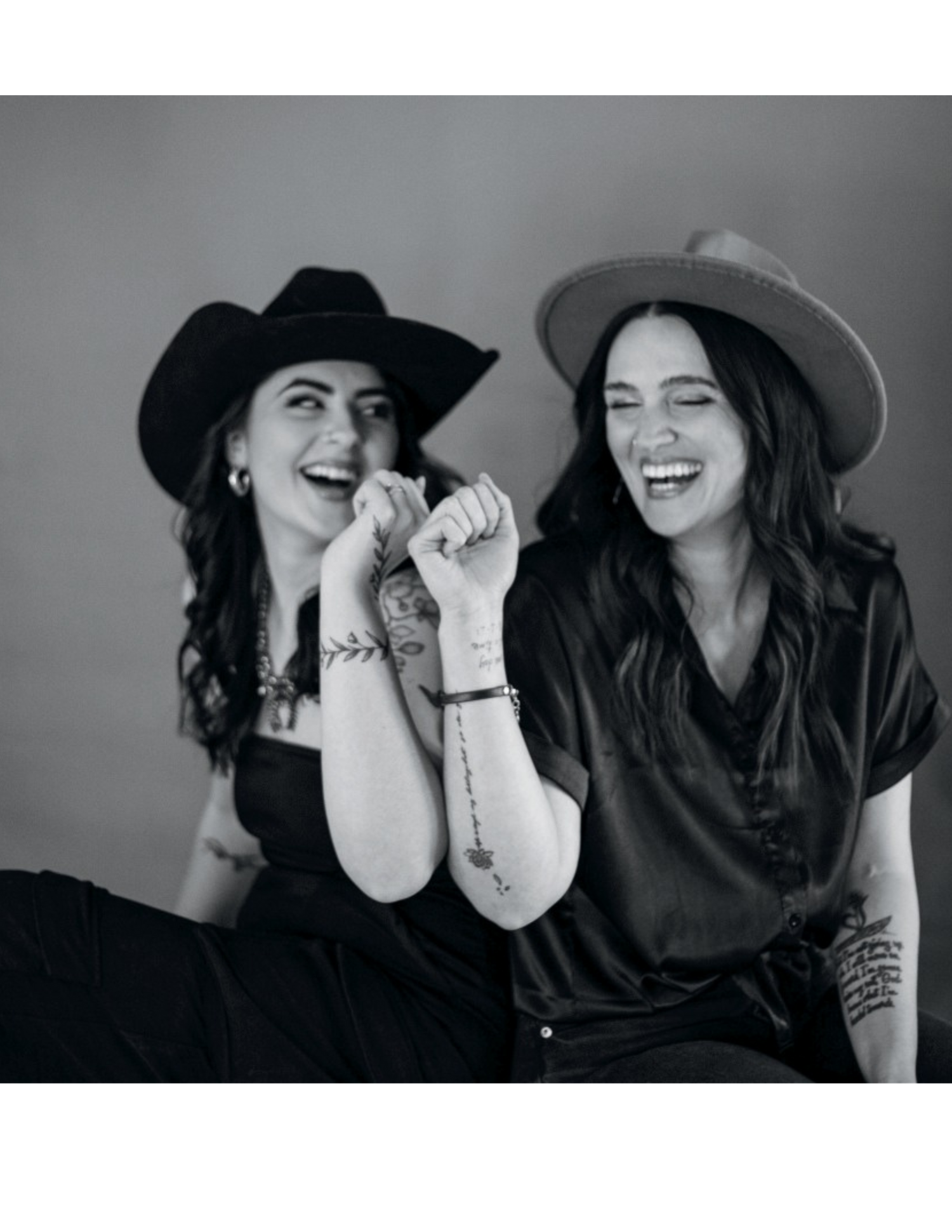 Black and white candid photo showing two women in hats laughing together while seated against plain background.