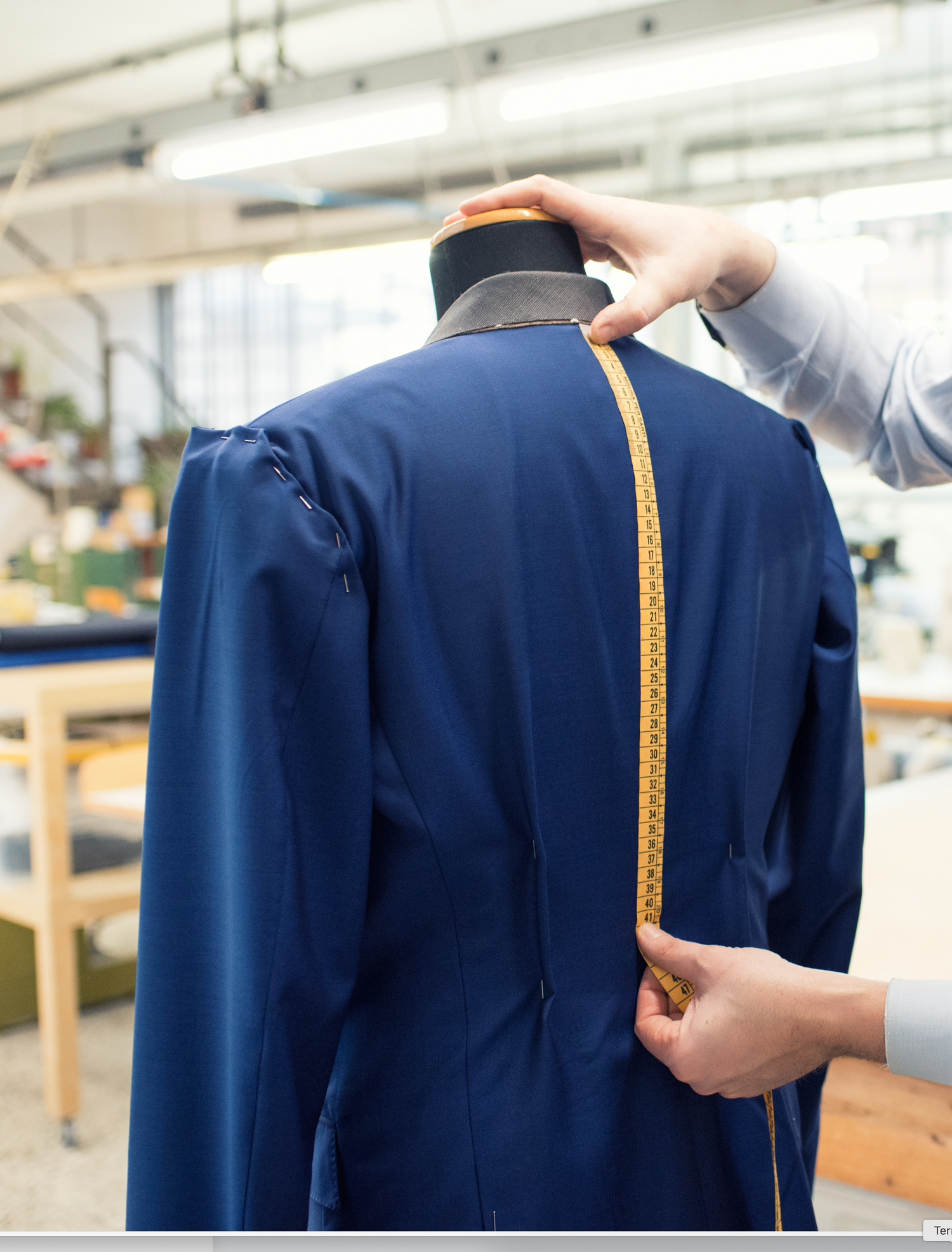 A tailor measuring the back length of a navy blue formal jacket with a measuring tape.