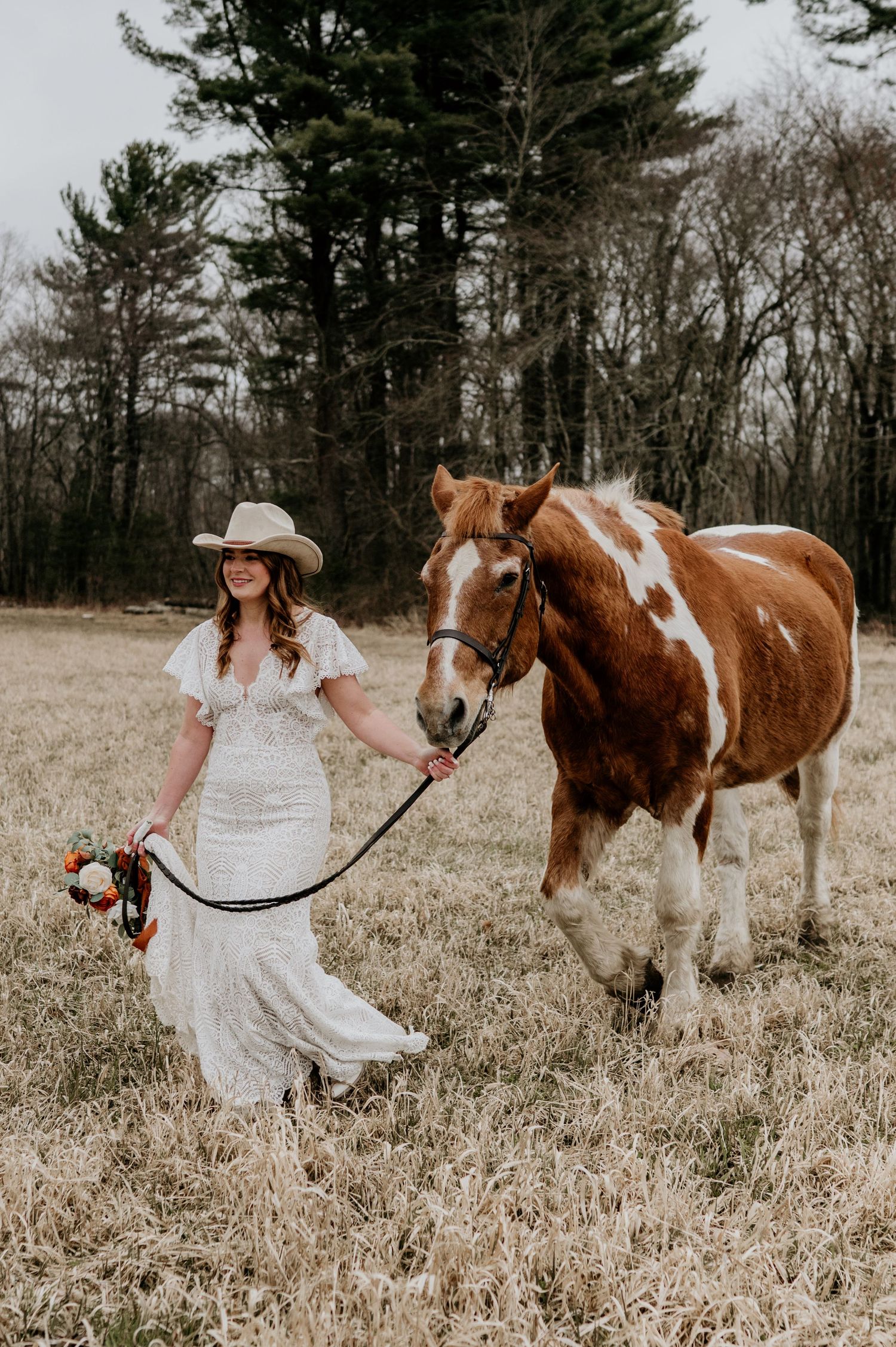 A rustic country bride in a white lace dress stands with her brown and white pinto horse in a winter field.