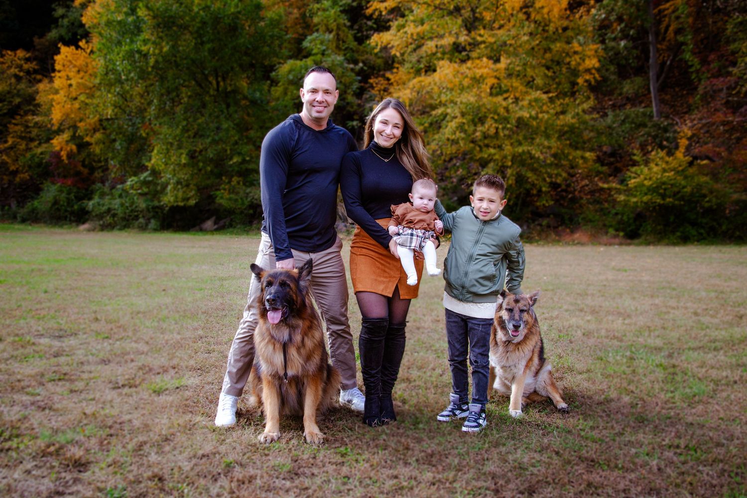 Family and dogs posing together in autumn field with fall foliage in background.