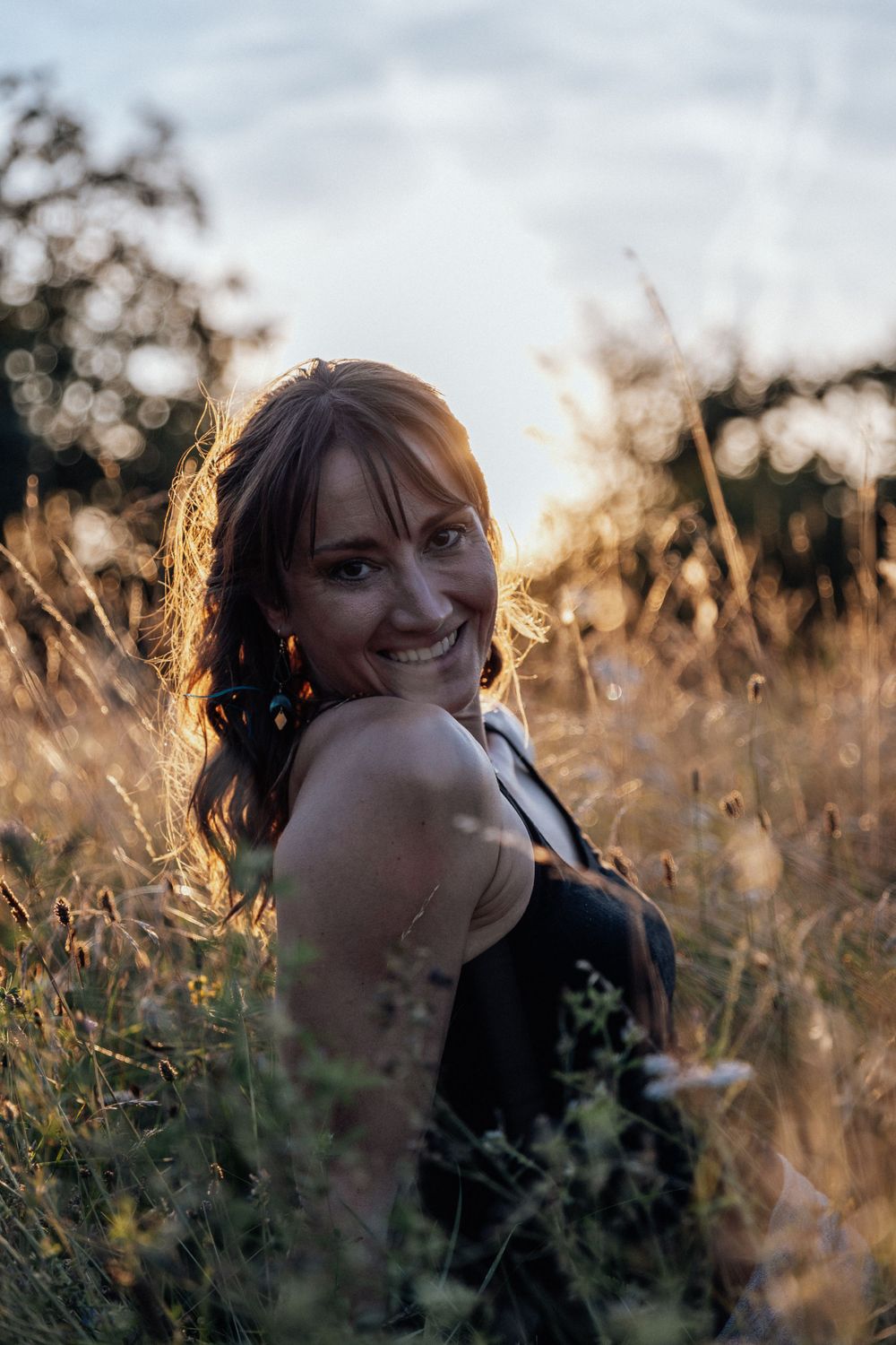 Golden sunlight illuminates a portrait of someone in a black tank top sitting among tall grass at sunset.