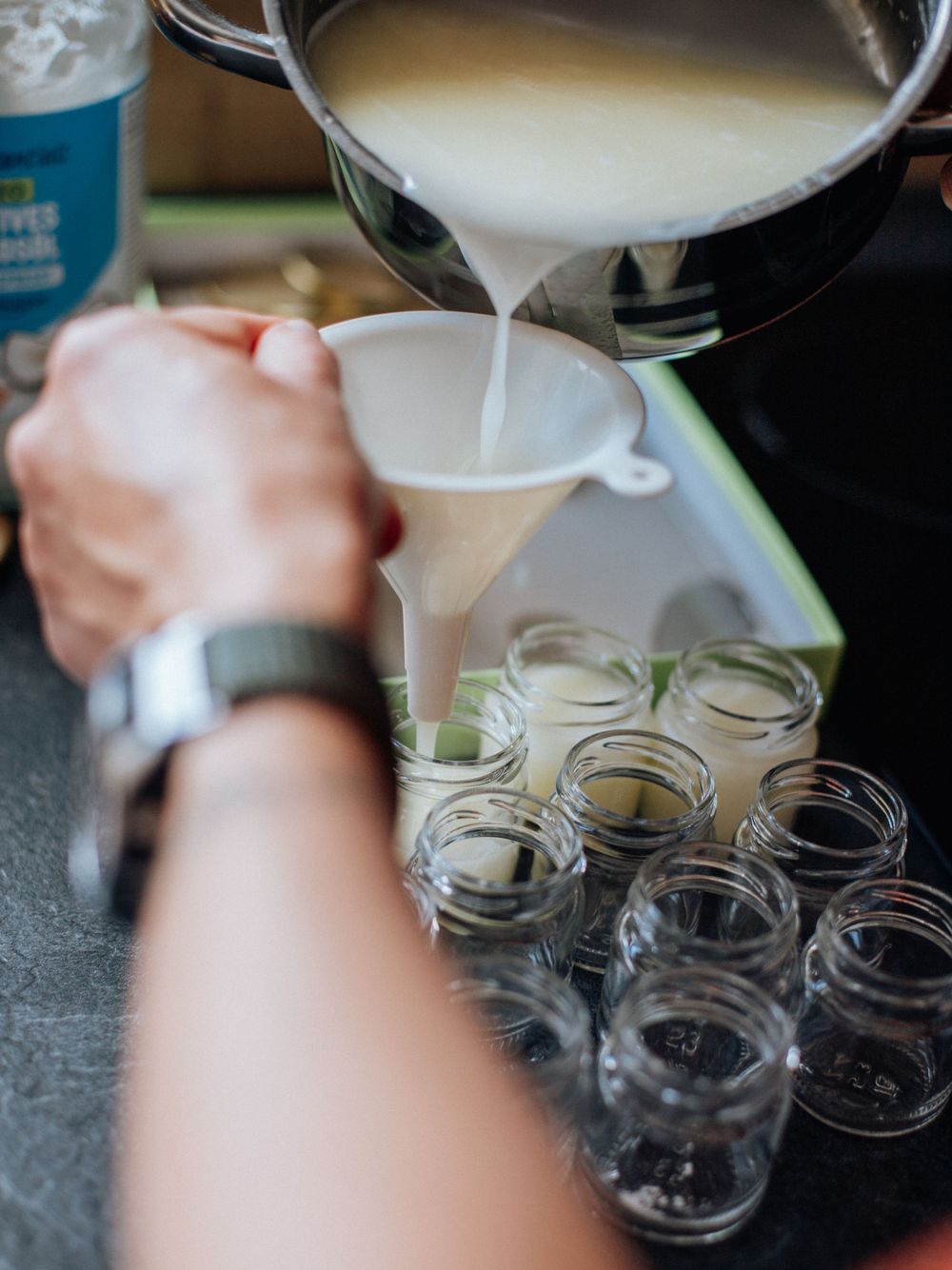 A white liquid mixture is being poured through a funnel into a row of empty glass mason jars on a dark countertop.