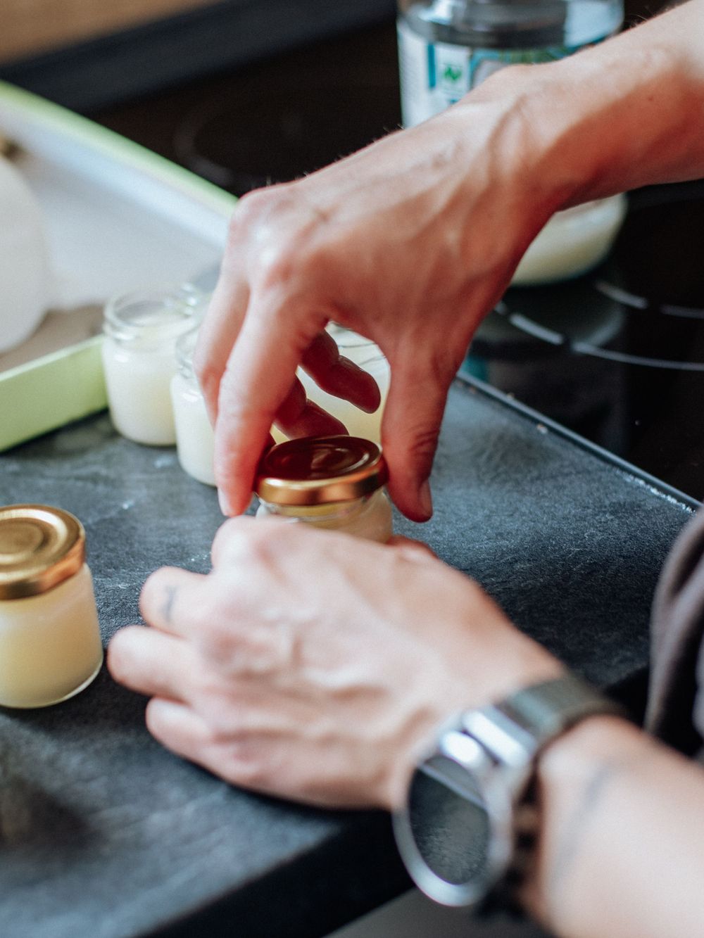 Hands prepare small glass jars on a dark surface with creamy ingredients for dessert making.