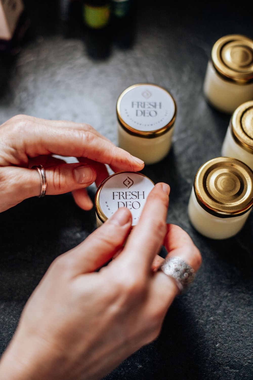 Hands with silver rings placing labels on glass jars of fresh homemade products on dark surface.