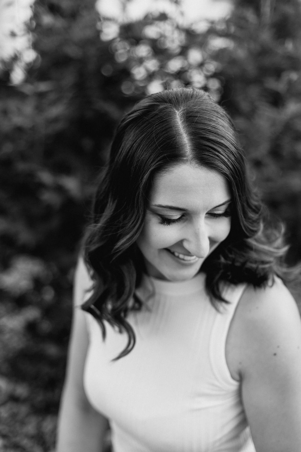 A person in a white sleeveless top smiles while looking down in a black and white portrait.