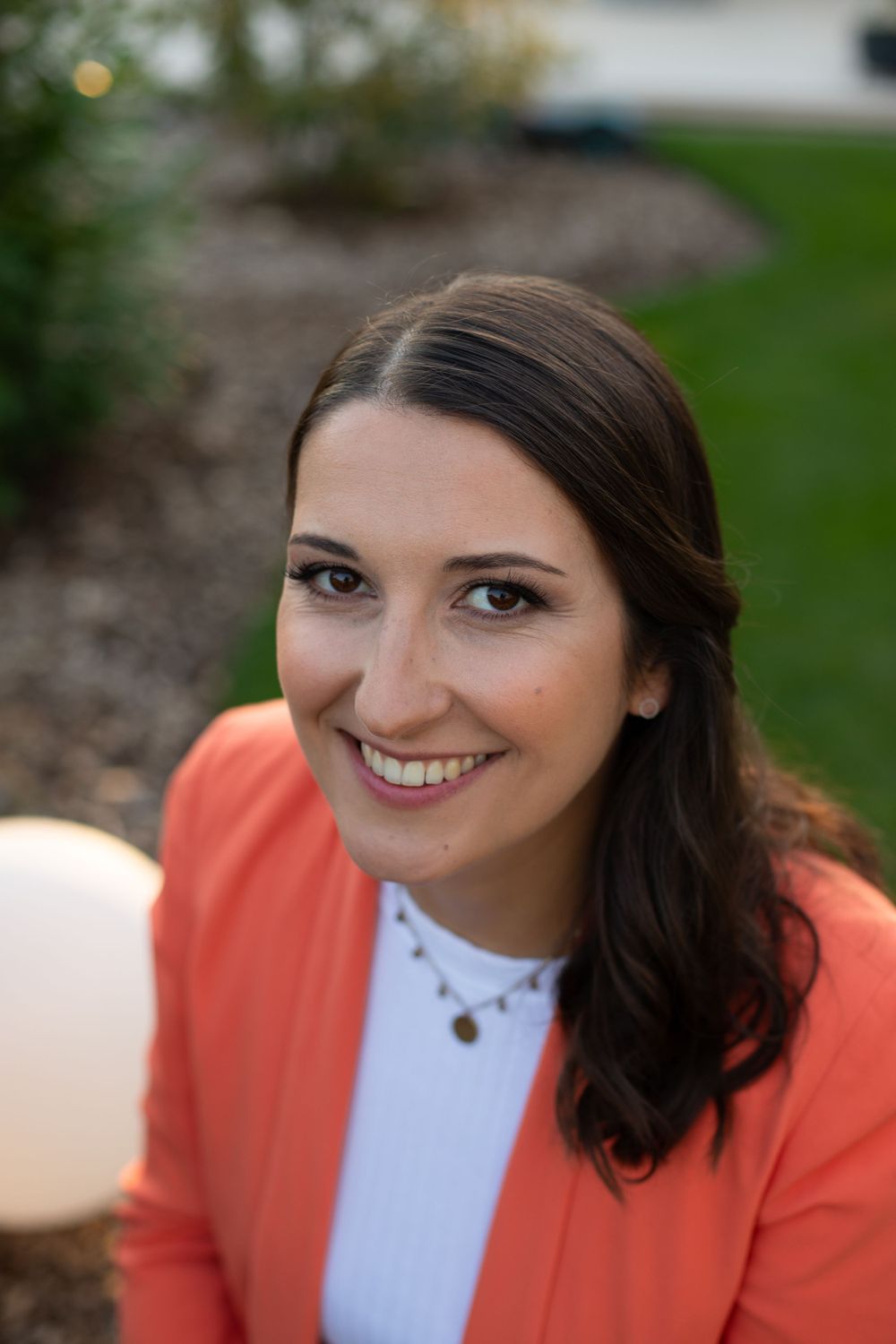 Professional headshot of someone wearing a coral blazer and white top smiling warmly against a blurred outdoor background.