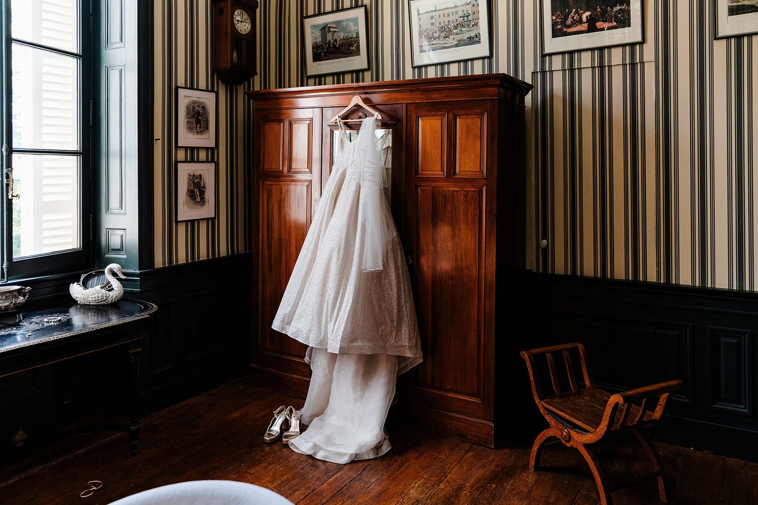 White wedding gown displayed on hanger against dark wood paneled wall with striped wallpaper.