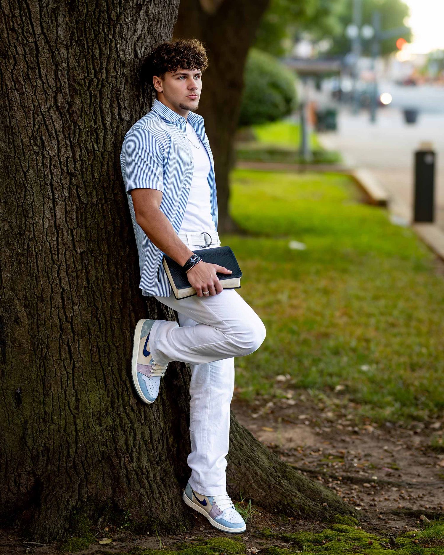 Casual portrait leaning against tree wearing blue shirt and white pants.