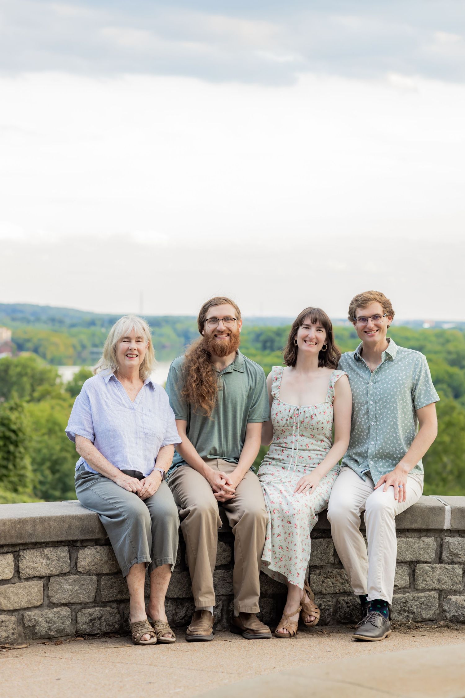 Four smiling people sitting on a stone wall outdoors with a scenic green landscape and cloudy sky in the background.