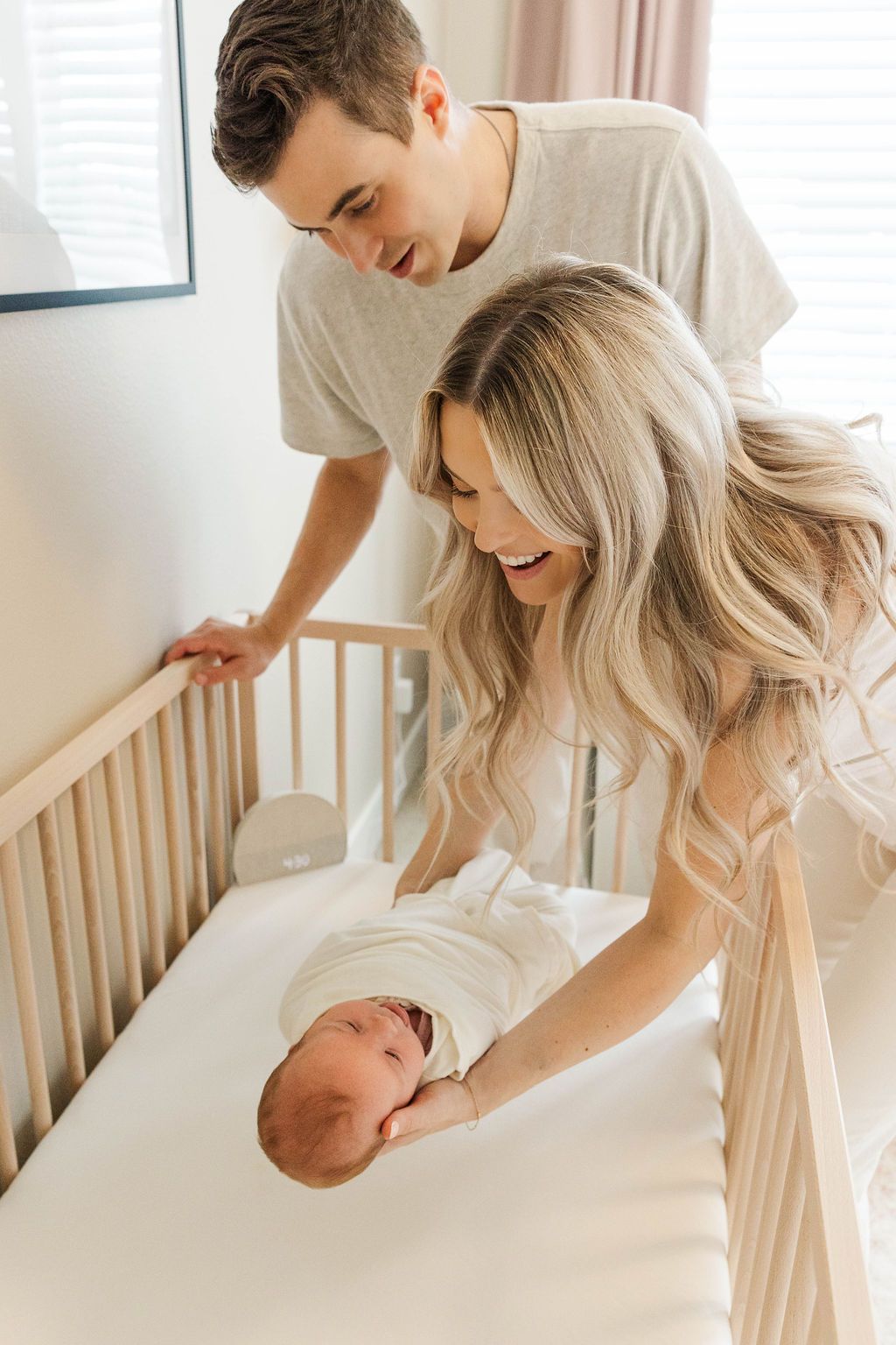 Happy parents smile at their swaddled newborn baby lying in a wooden crib.