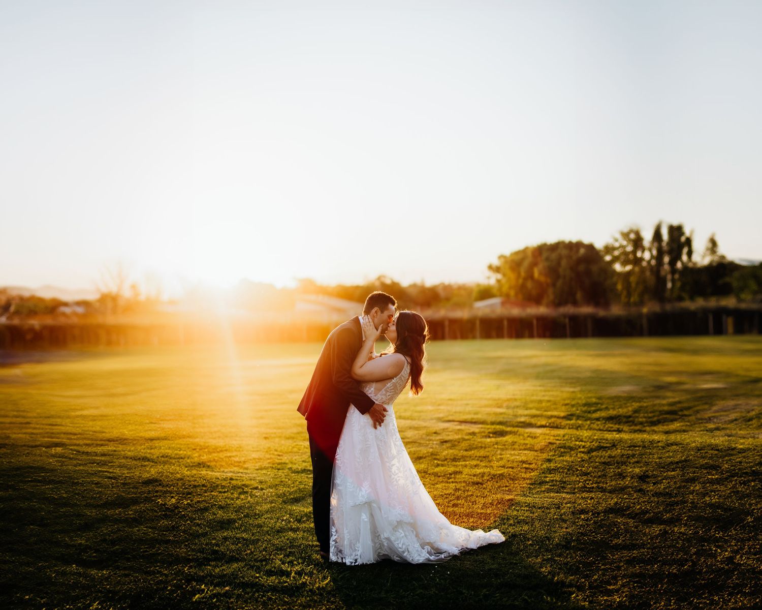 A romantic sunset wedding photo of a couple embracing on a grassy field with golden light streaming behind them.