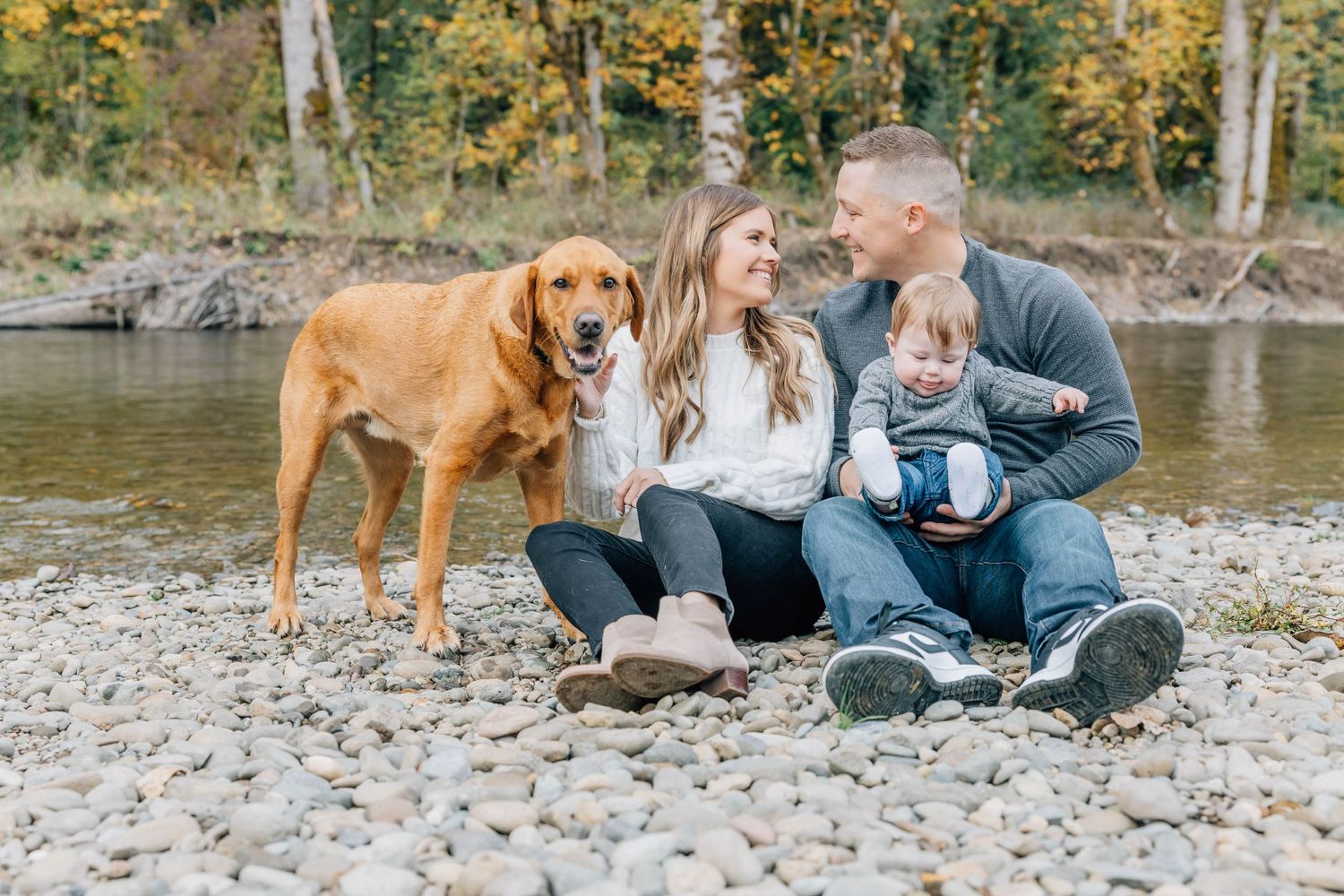 Family with golden retriever dog sitting on rocky riverbank surrounded by autumn foliage.