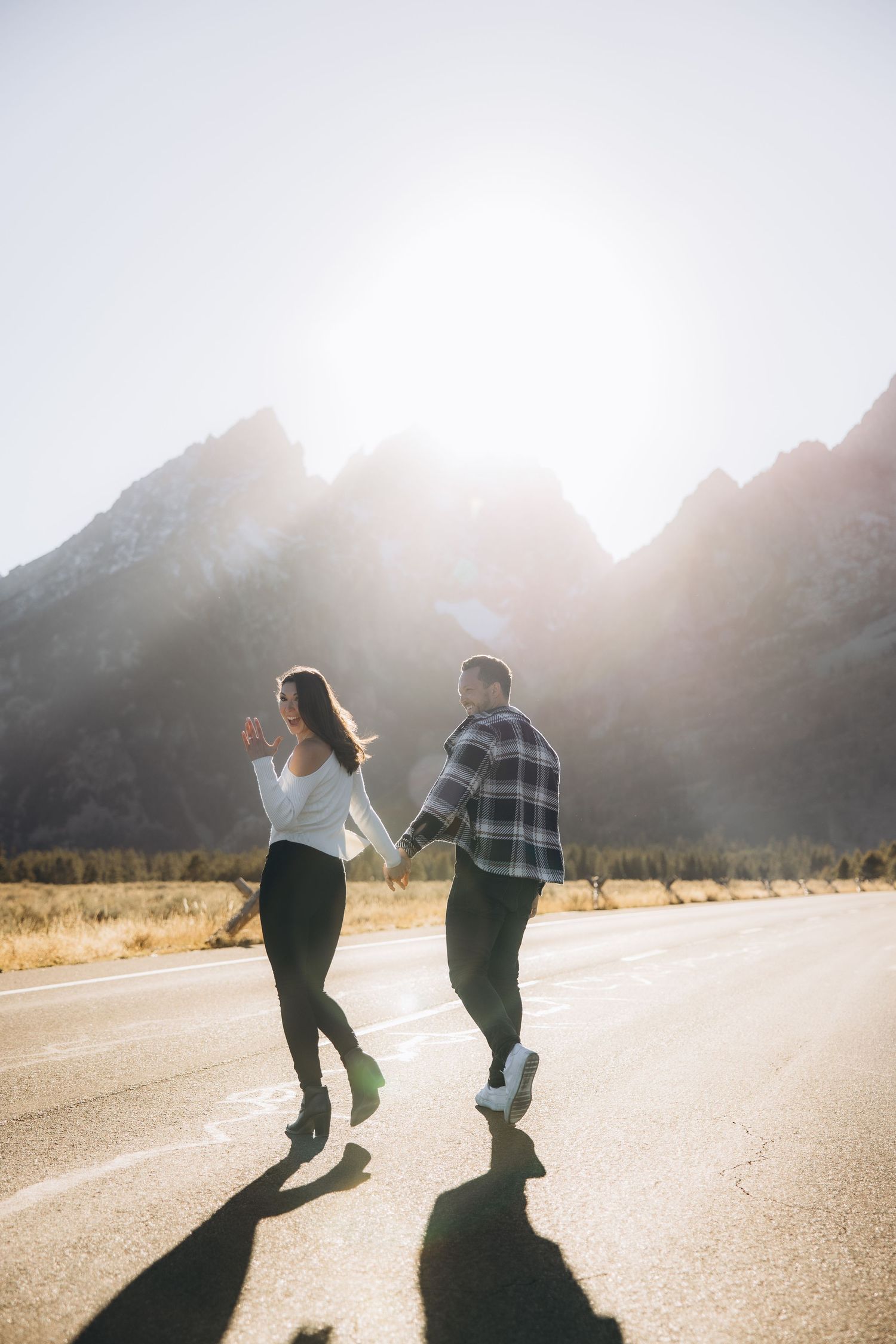 Two people holding hands while walking on an empty mountain road during sunset, casting long shadows on the pavement.