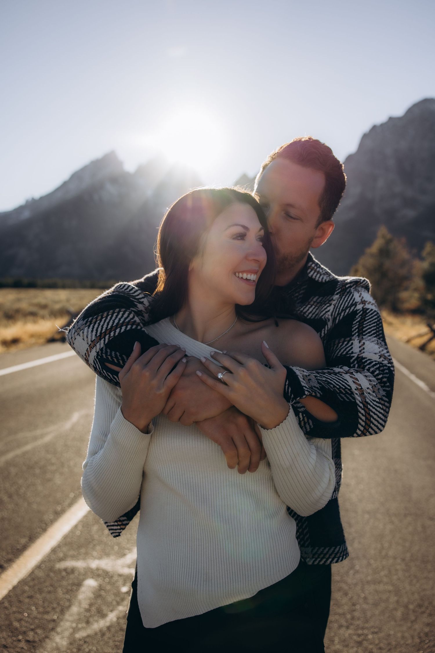 A romantic couple embraces on a mountain road with sunlight streaming through misty peaks in the background.