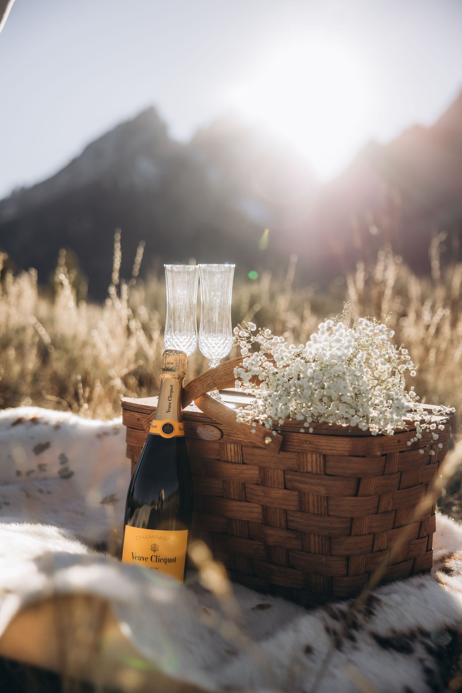 Champagne bottle and flutes nestled in woven basket with baby's breath flowers against snowy mountain backdrop at sunset.