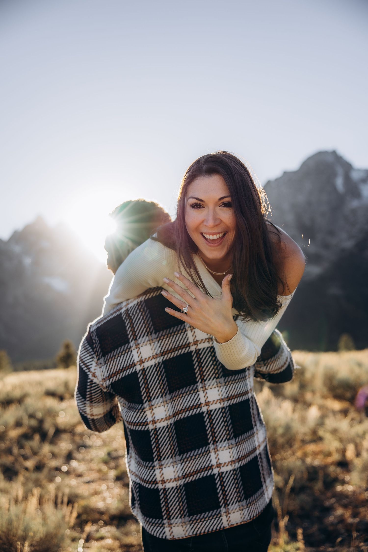 Two people share a joyful piggyback moment outdoors with mountains and sunlight in the background.