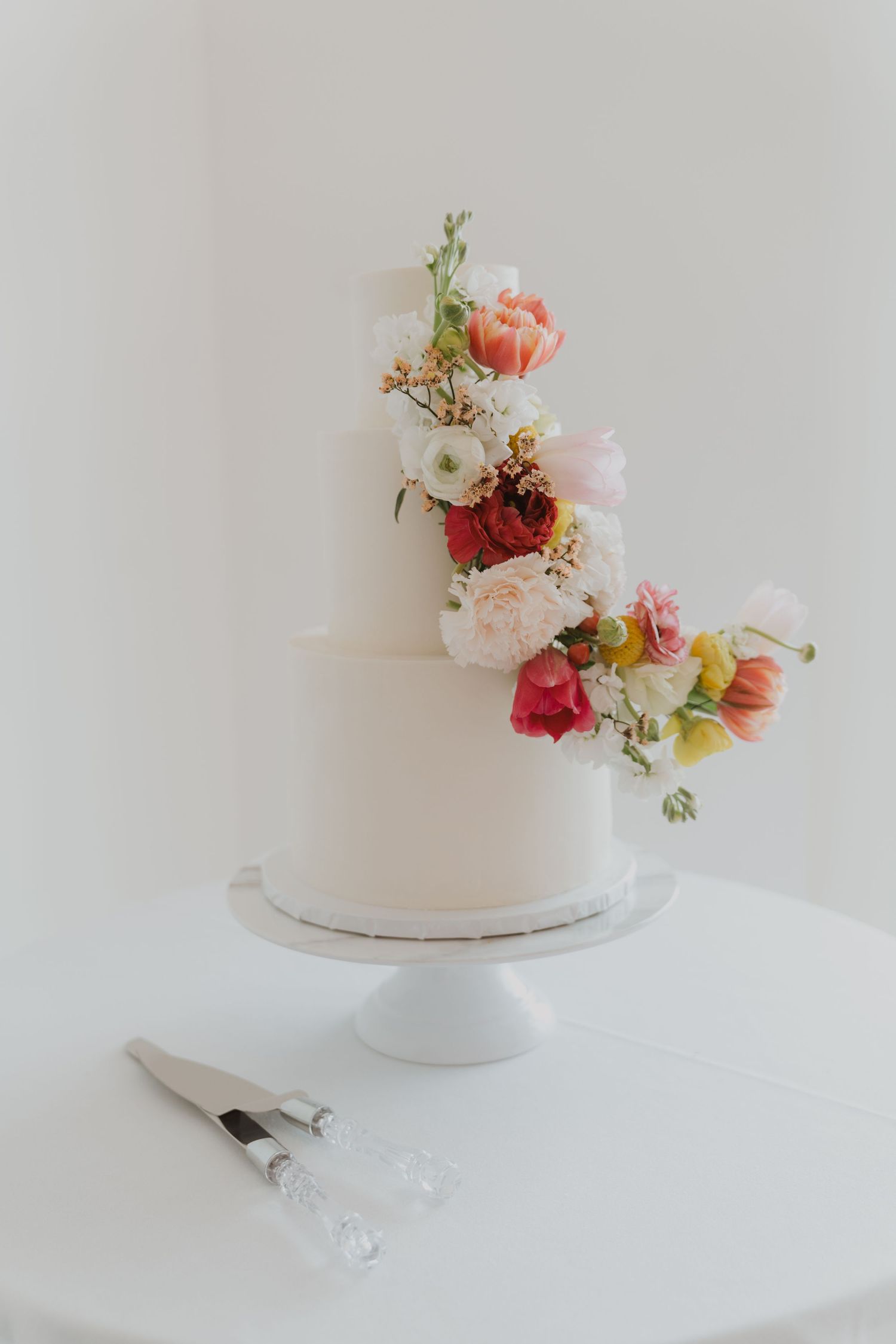 Elegant white wedding cake decorated with fresh pink and coral flowers on a white cake stand.