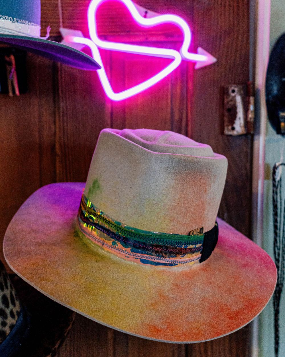 Western style hat displayed under pink neon heart sign on wooden wall.