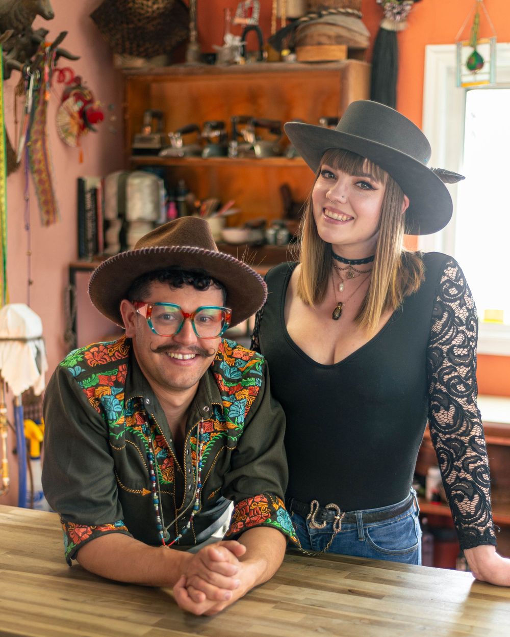 Stylish duo in western hats and vintage attire pose at rustic indoor setting.
