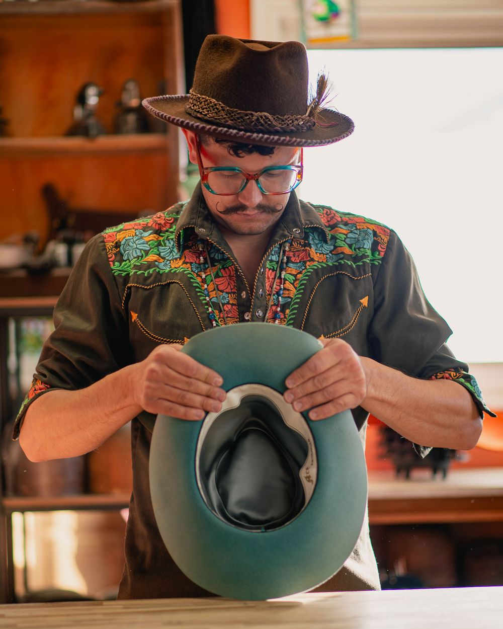 Artisan in western wear examines a turquoise cowboy hat while working in a hat shop.