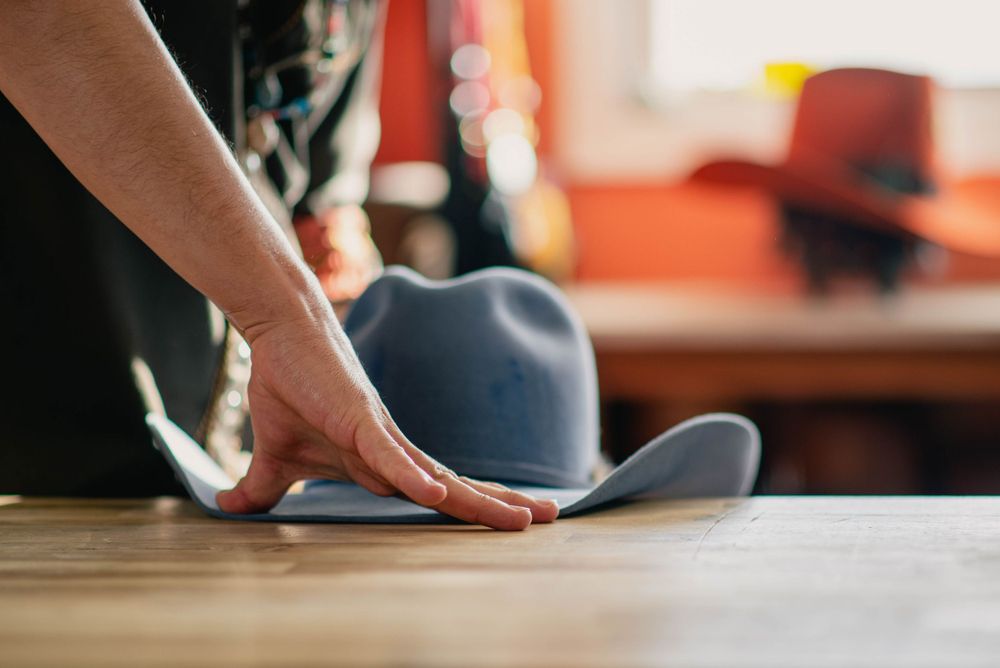 Hands carefully shaping gray felt hat on wooden workspace surface.