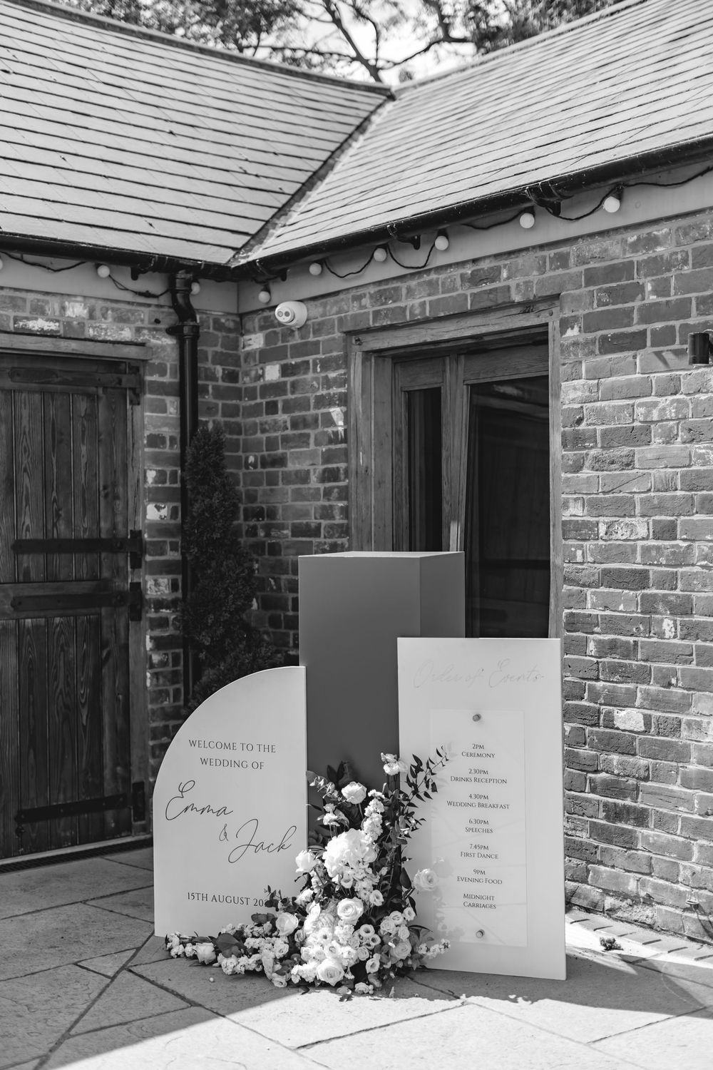 Black and white photo of wedding welcome sign with floral arrangement against brick wall.