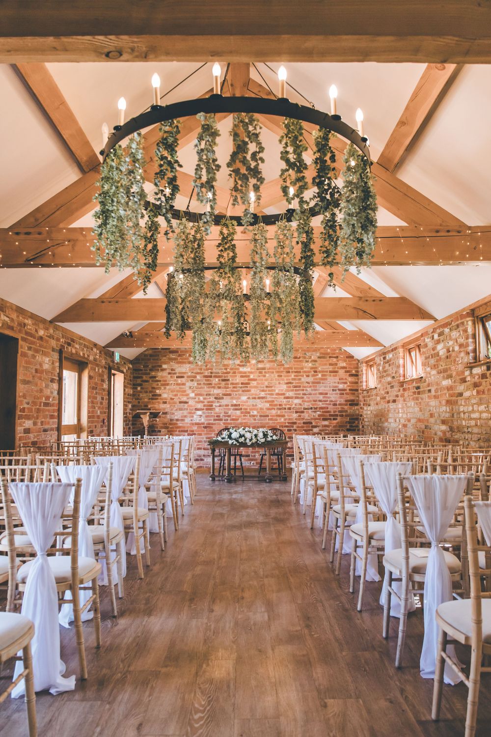 Rustic barn wedding venue with exposed beams, brick walls and white chairs decorated with hanging greenery.