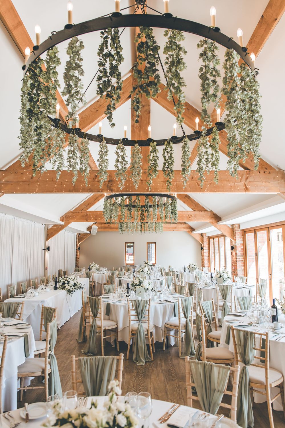 Elegant wedding reception venue with exposed wooden beams, hanging greenery, and white table settings.