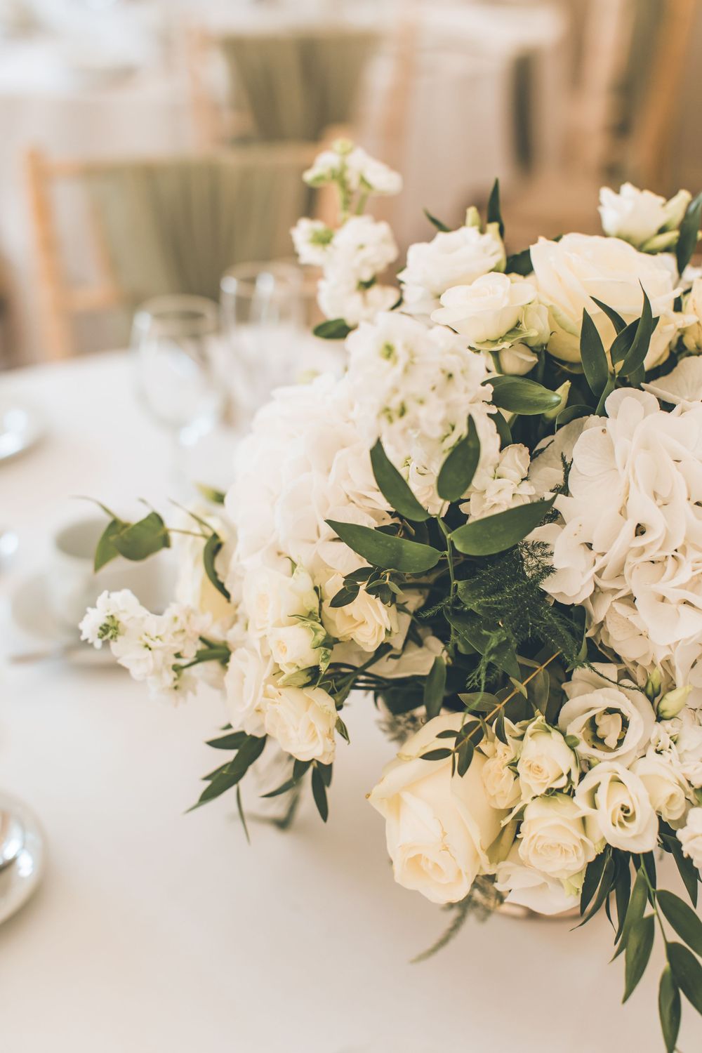 Elegant white floral centerpiece with roses and greenery on a white tablecloth at a wedding reception.