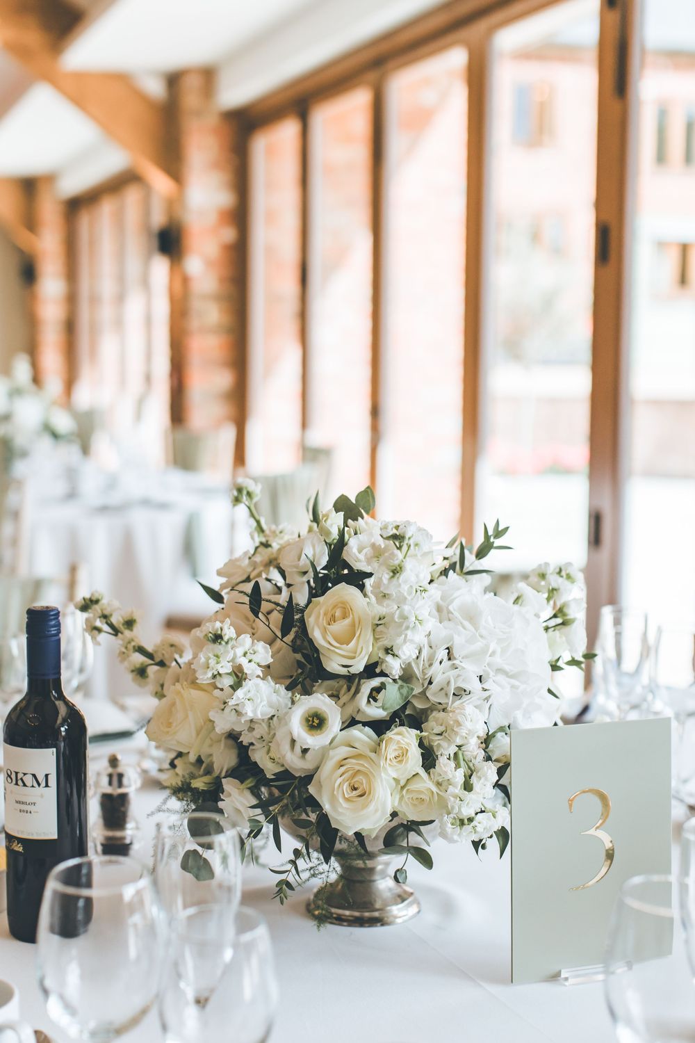 White floral centerpieces and wine bottles arranged on long tables with natural lighting.