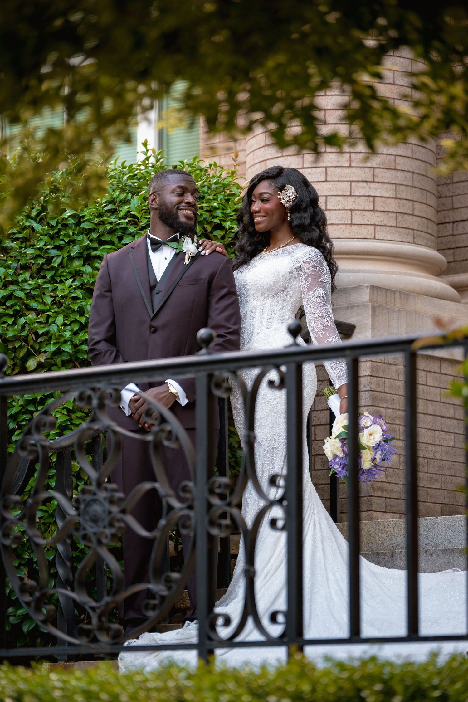 Elegant bride and groom in formal wedding attire pose on decorative iron staircase.