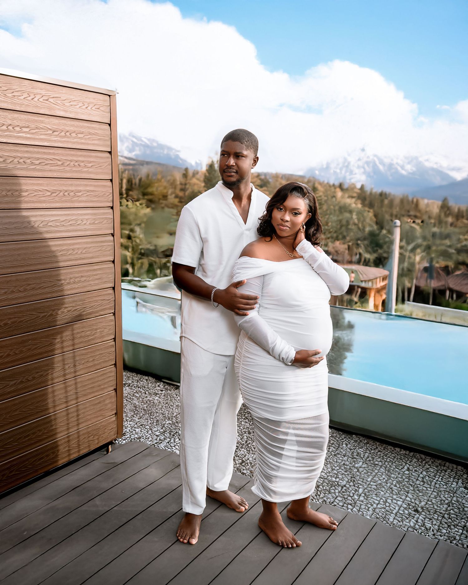 A couple in matching white outfits pose together on a deck with mountains and a pool in the background.