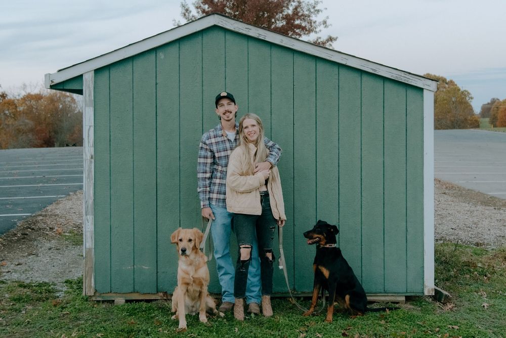 A couple poses with their two dogs in front of a mint green shed.